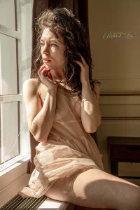 Volwassen vrouw poseert tijdens een artistieke naaktfotoshoot in een sfeervol interieur.